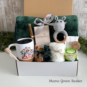 May include: A gift basket containing a dark green plush blanket, a white mug with a floral MAMA graphic, a wooden honey dipper, a glass straw, a Moonstruck chocolate bar, a gray crochet hippo rattle, a white washcloth, a green candle, and caramels.