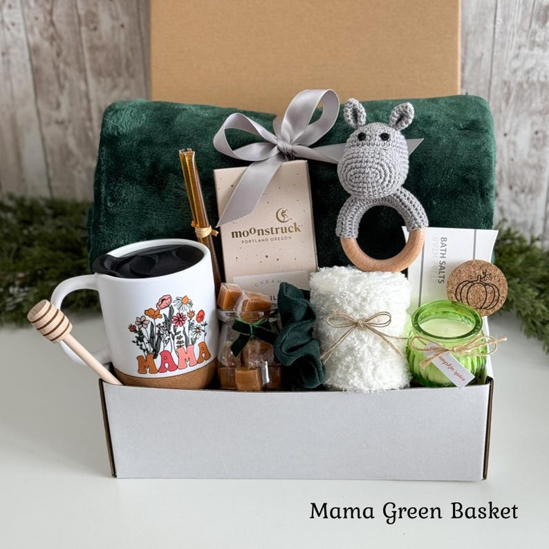 May include: A gift basket containing a dark green plush blanket, a white mug with a floral MAMA graphic, a wooden honey dipper, a glass straw, a Moonstruck chocolate bar, a gray crochet hippo rattle, a white washcloth, a green candle, and caramels.