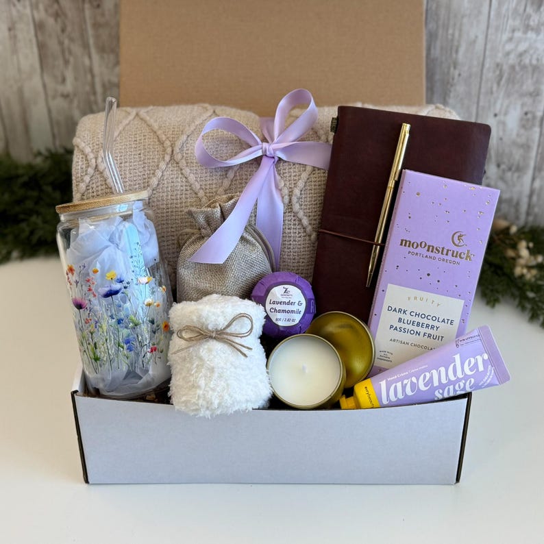 May include: A gift box containing a glass tumbler with a floral print, a beige knit blanket with a lavender ribbon, a small burlap pouch, a dark brown journal with a gold pen, a lavender bath bomb, a candle, and a lavender sage hand cream.