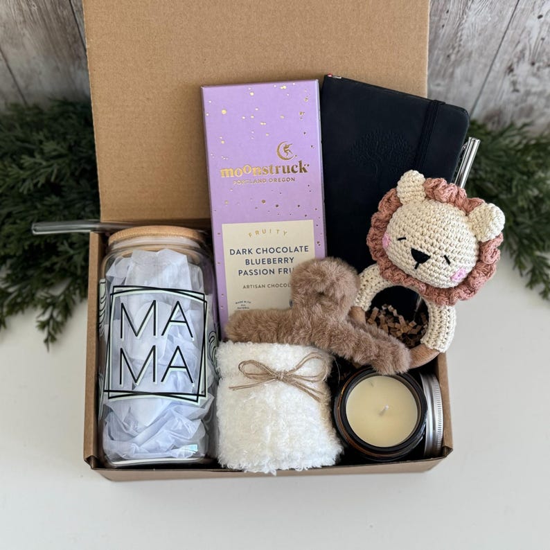 K&ouml;nnte beinhalten: Eine Geschenkbox mit einem Glastrinkbecher mit Bambusdeckel und der Aufschrift MAMA, einer Moonstruck Zartbitterschokolade, einem schwarzen Notizbuch, einer geh&auml;kelten L&ouml;wenrassel, einer flauschigen braunen Haarspange, einem wei&szlig;en Accessoire und einer Kerze.