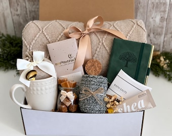 May include: A gift box containing a beige knit blanket with a ribbon, a white mug with a gold spoon, a Moonstruck chocolate bar, a jar of caramels, a gray knit item, a green journal with a tree icon, bath salts, and a shea butter tube.