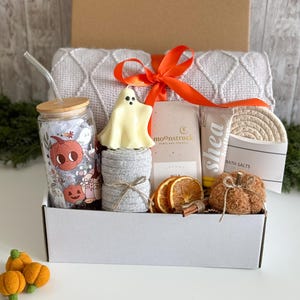 K&ouml;nnte beinhalten: Eine wei&szlig;e Geschenkbox mit herbstlichen Artikeln. Enth&auml;lt ein Glas mit K&uuml;rbisdesign, eine geisterf&ouml;rmige Kerze, eine graue Decke und einen kleinen k&uuml;rbisf&ouml;rmigen Gegenstand. Ein orangefarbenes Band bindet die Box zusammen.