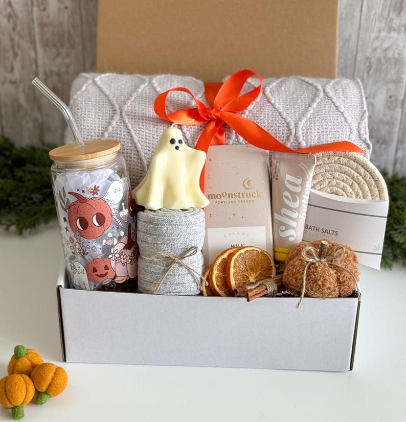 K&ouml;nnte beinhalten: Eine wei&szlig;e Geschenkbox mit herbstlichen Artikeln. Enth&auml;lt ein Glas mit K&uuml;rbisdesign, eine geisterf&ouml;rmige Kerze, eine graue Decke und einen kleinen k&uuml;rbisf&ouml;rmigen Gegenstand. Ein orangefarbenes Band bindet die Box zusammen.
