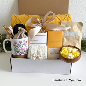 May include: A gift box containing a yellow textured blanket, a white mug with floral designs and the text MAMA love cherish care, a wooden honey dipper, a small pouch, bath salts, a Moonstruck milk chocolate bar, and a succulent-shaped candle.