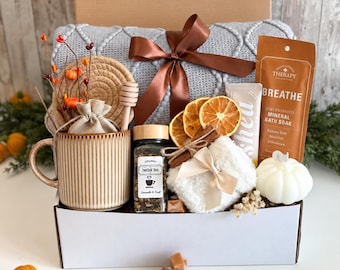 May include: A gift box containing a gray knit blanket with a brown ribbon, a ribbed ceramic mug, a jar of herbal tea, a wooden honey dipper, dried orange slices, a bath soak packet labeled BREATHE, a white pumpkin candle, and a small pouch.