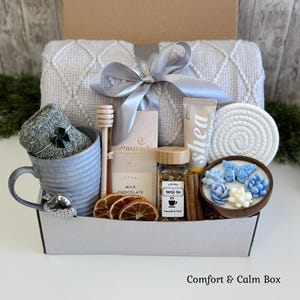 May include: A gift box containing a grey textured throw blanket with a silver ribbon, a grey mug with a heart-shaped tea infuser, a honey dipper, a Moonstruck milk chocolate bar, shea cream, herbal tea, cinnamon sticks, dried orange slices, and a candle.