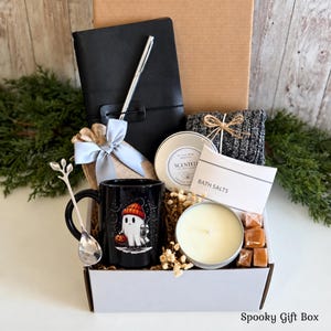 May include: A gift box containing a black notebook with a silver pen, a black mug featuring a ghost graphic, a silver spoon, a scented candle in a tin, a bag of bath salts, a pair of grey knit socks, and small caramel-colored cubes.