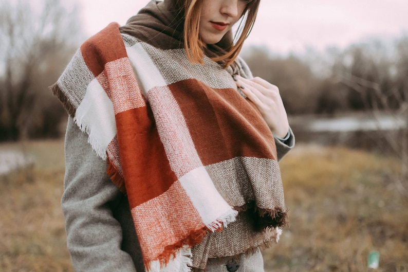 May include: A woman wearing a brown, white, and red plaid scarf with frayed edges. The scarf is wrapped around her neck and draped over her shoulders.