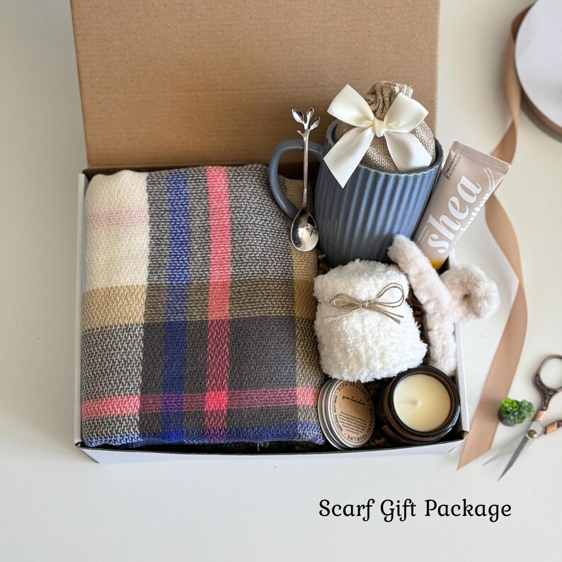 May include: A gift box containing a plaid scarf with gray, blue, and pink tones, a blue ribbed mug with a white bow, a silver spoon, a tube of shea cream, a white fuzzy accessory, and a small candle. The text Scarf Gift Package is visible.