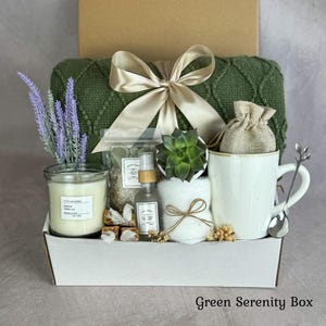 May include: A gift set in a white box containing a dark green knitted blanket with a beige ribbon, a vanilla candle, a small succulent, a spray bottle, a white mug with a silver spoon, a burlap pouch, and dried lavender sprigs.
