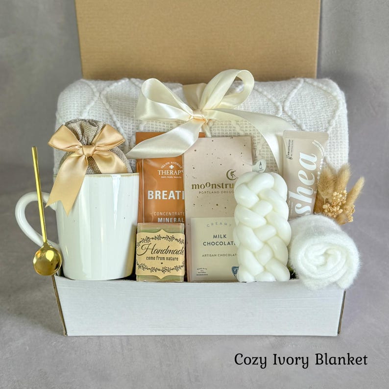 Pu&ograve; includere: Una confezione regalo contenente una tazza in ceramica bianca con cucchiaino dorato, una coperta in maglia color crema con fiocco in raso, una saponetta artigianale, una tavoletta di cioccolato al latte Moonstruck, burro di karit&eacute; e una candela.