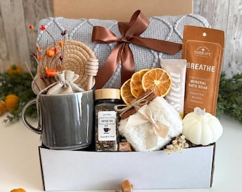 May include: A gift box containing a gray ceramic mug, a jar of herbal tea, a brown ribbon-tied gray knit blanket, a bag of mineral bath soak labeled BREATHE, dried orange slices, a white pumpkin candle, and a small wooden honey dipper.