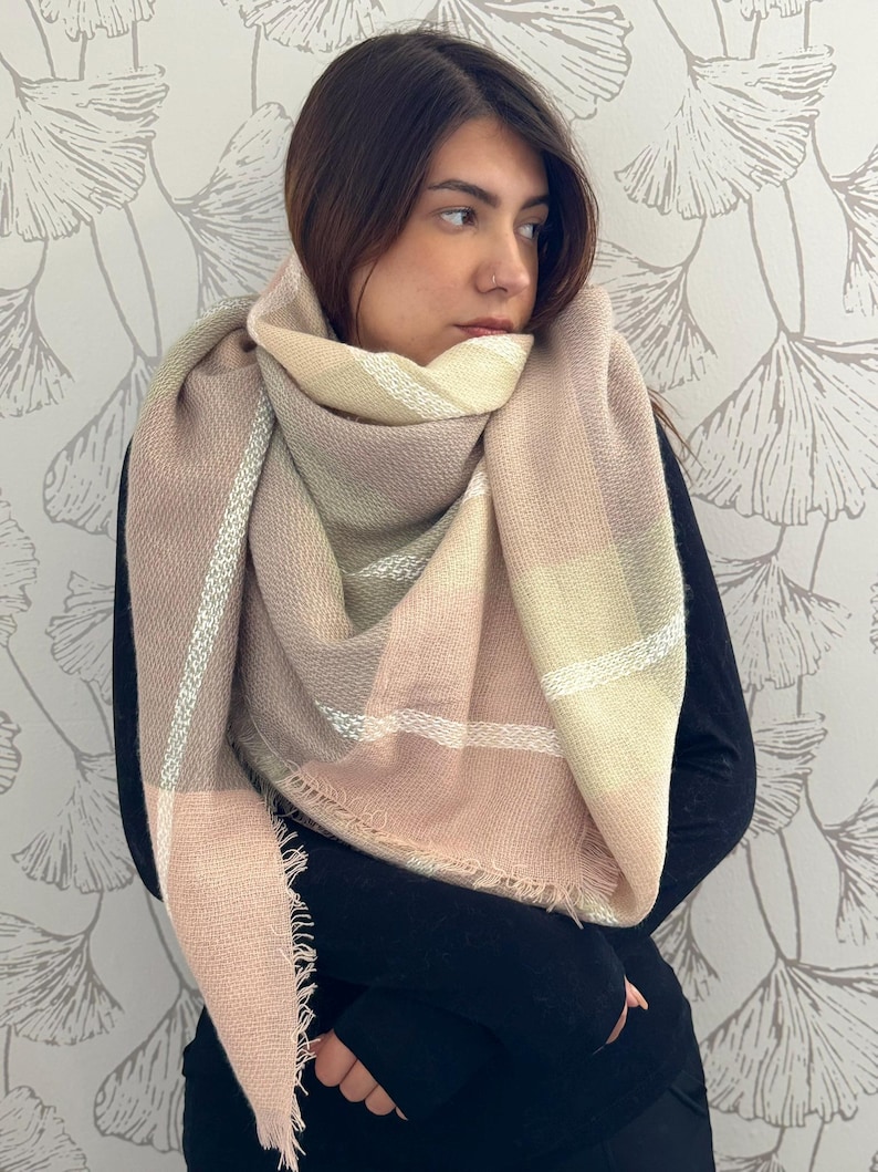 May include: A large, triangular scarf featuring a soft plaid pattern in muted shades of dusty rose, cream, beige, and sage green. The fabric has a textured weave and is finished with frayed edges along the bottom, draped loosely around the neck.