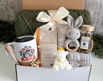 May include: A gift box containing a dark green textured blanket, a white mug with a floral MAMA graphic, a honey dipper, an amber glass straw, a Moonstruck milk chocolate bar, a shea cream tube, a crochet bunny rattle, and herbal tea.