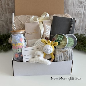 May include: A gift box containing a glass tumbler with a straw and floral text, a Moonstruck chocolate bar, a shea cream tube, a dark gray notebook, a succulent candle, a crochet giraffe toy, a white hair claw, and a gray striped fabric item.