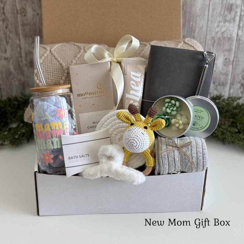 May include: A gift box containing a glass tumbler with a straw and floral text, a Moonstruck chocolate bar, a shea cream tube, a dark gray notebook, a succulent candle, a crochet giraffe toy, a white hair claw, and a gray striped fabric item.