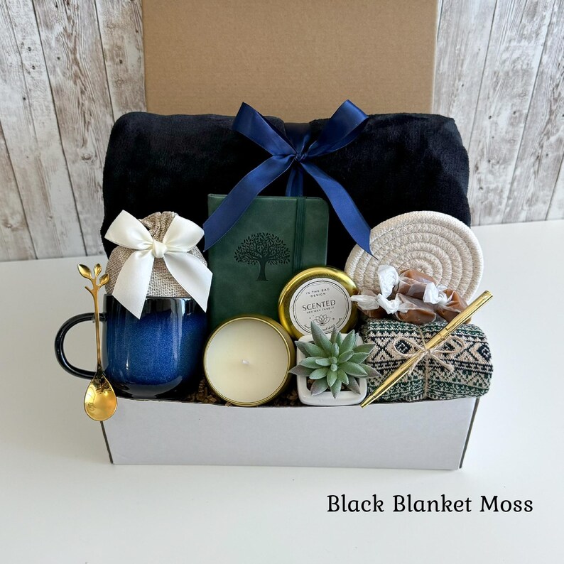 May include: A gift box containing a black blanket tied with a navy blue ribbon, a blue ceramic mug with a white bow, a green journal with a tree design, a gold-toned spoon, a scented candle, a small succulent, a coaster, and a gold pen.