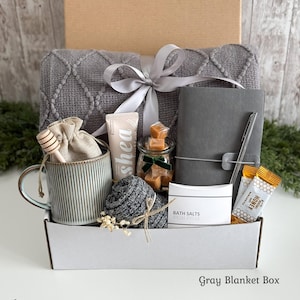 May include: A gift box containing a gray textured blanket tied with a silver ribbon, a ribbed ceramic mug with a wooden honey dipper, a tube of shea lotion, a jar of caramels, a gray notebook with a pen, bath salts, and honey packets.
