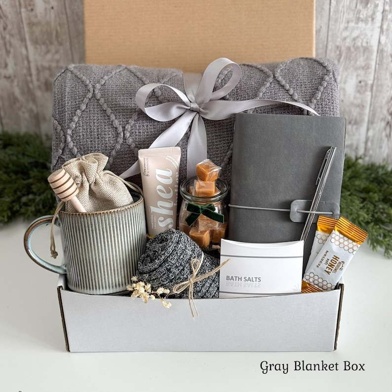 May include: A gift box containing a gray textured blanket tied with a silver ribbon, a ribbed ceramic mug with a wooden honey dipper, a tube of shea lotion, a jar of caramels, a gray notebook with a pen, bath salts, and honey packets.