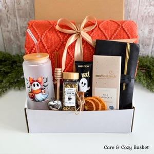 May include: A gift box containing an orange textured blanket with a gold ribbon, a frosted glass tumbler with a ghost graphic, a wooden honey dipper, a tea jar, a tube of hand cream, a Moonstruck milk chocolate bar, and a black notebook with a pen.