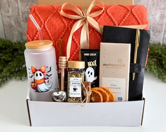 May include: A gift box containing an orange textured blanket with a beige ribbon, a frosted glass tumbler with a ghost graphic, a wooden honey dipper, a jar of herbal tea, a tube of hand cream, a Moonstruck milk chocolate bar, and a black notebook with a gold pen.