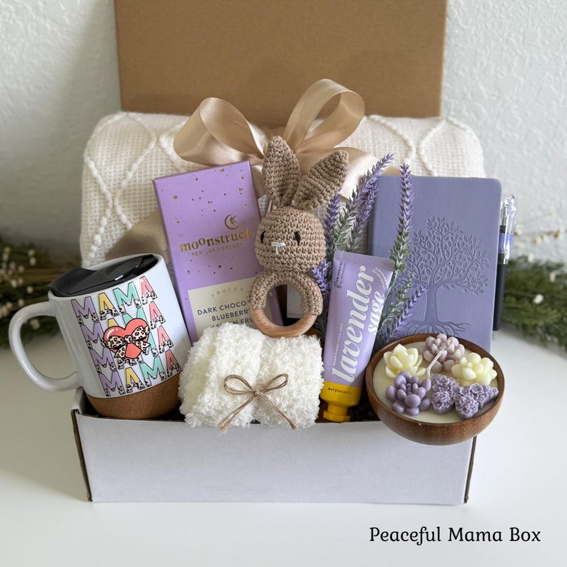K&ouml;nnte beinhalten: Eine Geschenkbox mit einer wei&szlig;en Tasse mit Korkboden und wiederholtem Mama-Text, einer lila Moonstruck-Schokolade, einer geh&auml;kelten Hasenrassel, einem wei&szlig;en flauschigen Tuch, einer Lavendel-Salbei-Tube, einem lila Notizbuch und einer Holzschale.