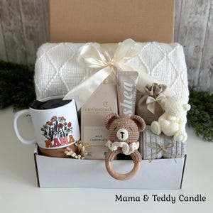 May include: A gift box containing a white ceramic mug with a floral Mama graphic, a Moonstruck milk chocolate bar, a tube of shea butter, a brown crocheted bear rattle, a small burlap pouch, a white bear-shaped candle, and a cream knit blanket.