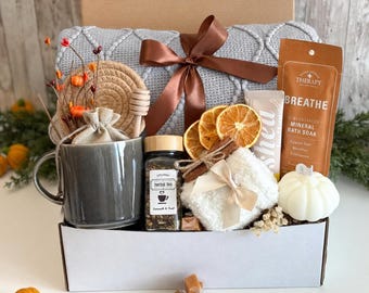 Può includere: Una scatola regalo contenente una tazza in ceramica grigia, un barattolo di tisana, una coperta in maglia grigia legata con un nastro marrone, un sacchetto di sali da bagno minerali BREATHE, fette d'arancia essiccate, una candela a zucca e un mestolo.