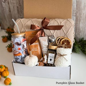 May include: A gift box containing a glass tumbler with a cow and pumpkin print, a metal straw, a bag of mineral bath soak, a shea cream tube, a heart-shaped tea infuser, a jar of herbal tea, and a white fuzzy mug wrap with dried orange slices.