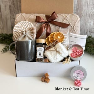 May include: A gift box containing a beige knit blanket with a brown ribbon, a gray ceramic mug, a wooden honey dipper, a jar of herbal tea, dried orange slices, a cinnamon stick, a white fuzzy pouch, bath salts, and a candle with pink floral wax.