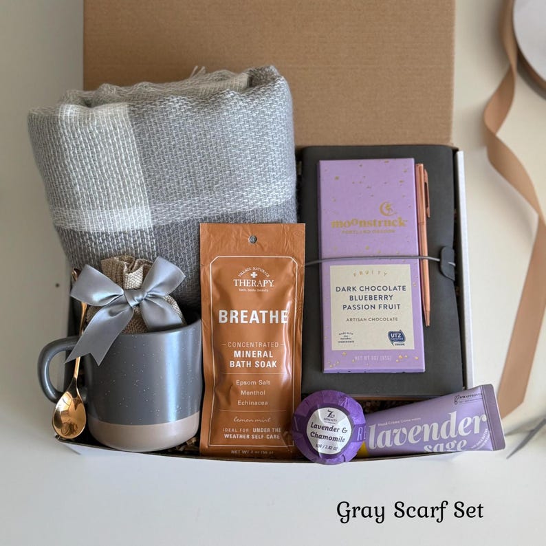 May include: A gift box containing a gray and white plaid scarf, a gray ceramic mug with a gold spoon and gray ribbon, a brown packet of Therapy Breathe mineral bath soak, a Moonstruck dark chocolate bar, a lavender bath bomb, and a lavender tube.
