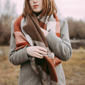 May include: A woman wearing a gray coat with a brown, red, and white plaid scarf. The scarf is wrapped around her neck and tied in a knot at the front.