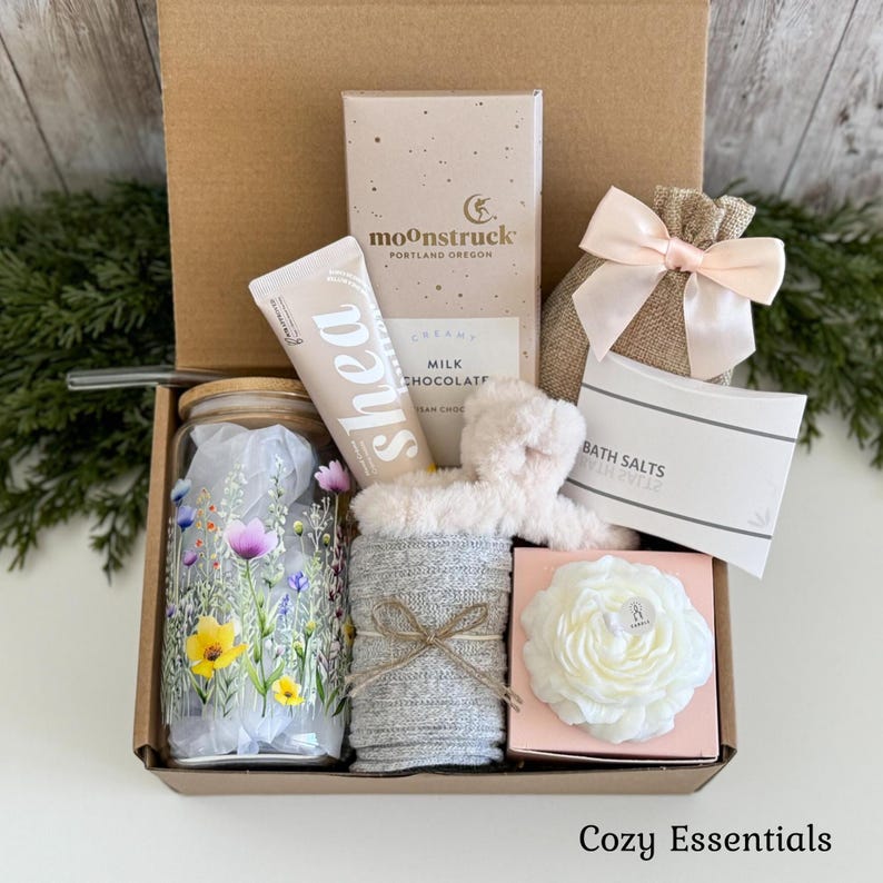 May include: A gift box containing a glass tumbler with a floral print, a tube of shea cream, a Moonstruck milk chocolate bar, a burlap bag with a pale pink bow, bath salts, a fuzzy headband, grey socks, and a white flower-shaped candle.