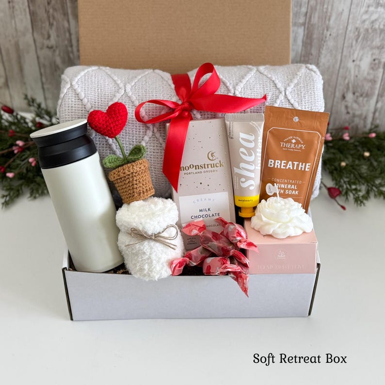 May include: A gift box containing a white tumbler, a crocheted heart plant, a white textured blanket with a red ribbon, a Moonstruck milk chocolate bar, a tube of shea butter, a Breathe mineral bath soak packet, and a white flower-shaped candle.