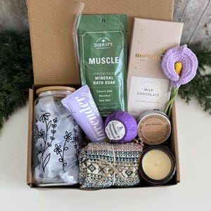 May include: A gift box containing a glass tumbler with a floral print, a clear glass straw, a green packet of muscle mineral bath soak, a lavender hand cream, a purple bath bomb, a Moonstruck milk chocolate bar, a crochet flower, and a candle.