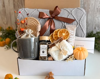 Può includere: Una confezione regalo contenente una coperta grigia testurizzata con un nastro marrone, una tazza in ceramica grigia, un barattolo di tisana, fette di arancia essiccata, una piccola pochette bianca soffice, un tubo di burro di karité, sali da bagno e una candela a forma di zucca.