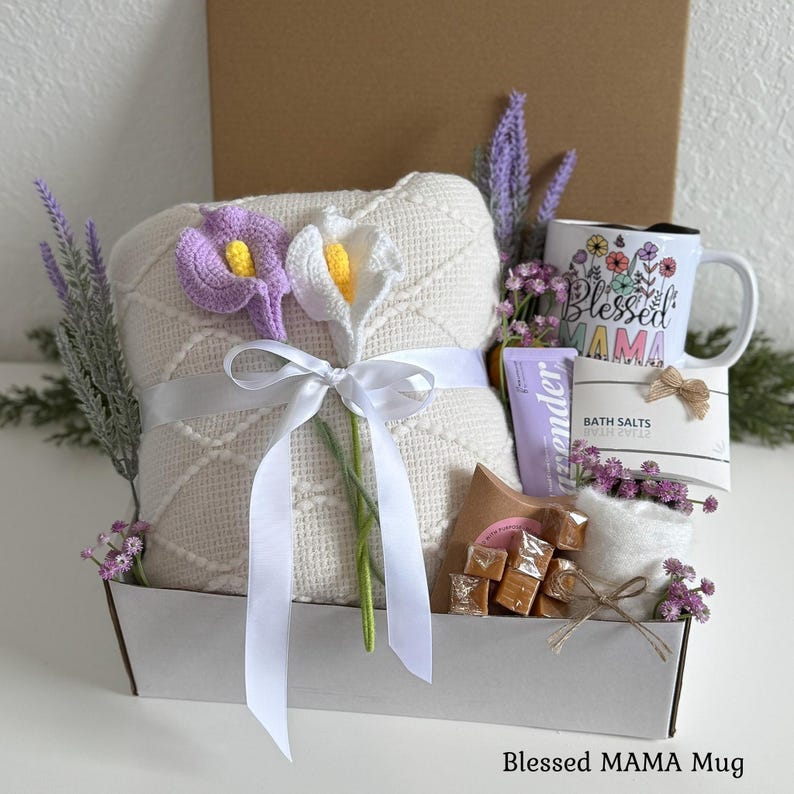 May include: A gift set containing a cream-colored textured blanket tied with a white ribbon and two crocheted calla lilies, a white mug with floral designs and the text Blessed MAMA, a tube of lavender lotion, bath salts, and square caramels.