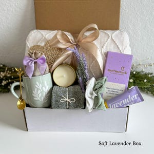 May include: A gift box containing a light green mug with white leaf patterns, a gold spoon, a candle in a gold tin, a grey knit item, a lavender-colored chocolate bar, a lavender tube, and a white textured blanket tied with a beige ribbon.