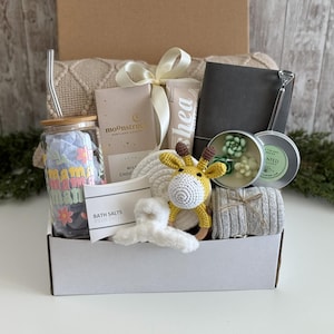 May include: A gift box containing a glass tumbler with a bamboo lid and straw, a Moonstruck chocolate bar, a shea cream tube, a gray notebook, a succulent candle, a crochet giraffe rattle, a white hair claw, bath salts, and a gray knit blanket.