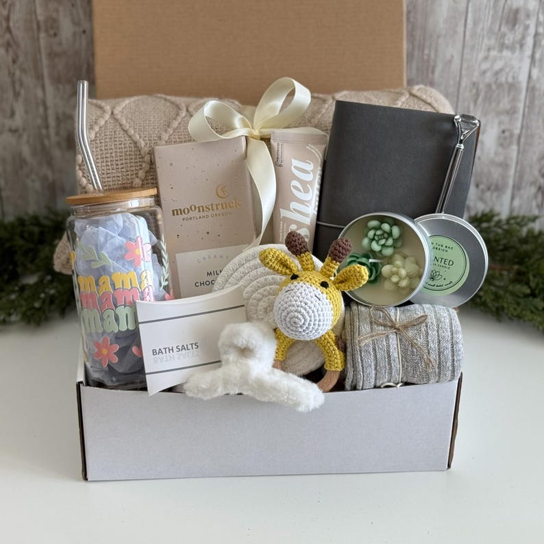 May include: A gift box containing a glass tumbler with a bamboo lid and straw, a Moonstruck chocolate bar, a shea cream tube, a gray notebook, a succulent candle, a crochet giraffe rattle, a white hair claw, bath salts, and a gray knit blanket.