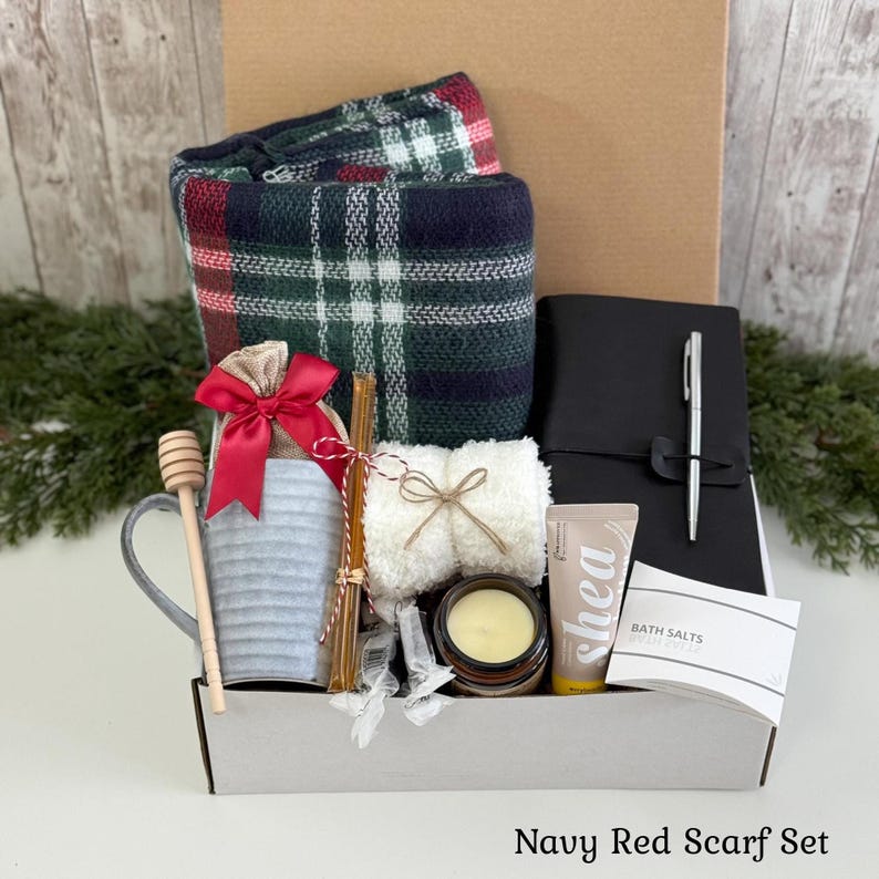 May include: A gift box containing a navy, red, and white plaid scarf, a black journal with a silver pen, a grey mug with a wooden honey dipper, a white fuzzy item, a candle, a tube of shea cream, and a packet labeled Bath Salts.