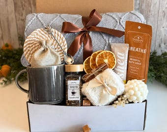 May include: A gift box containing a grey ceramic mug, a jar of herbal tea, a grey knit blanket with a brown ribbon, a bag of mineral bath soak, shea butter, dried orange slices, a white bubble candle, and a small linen pouch.