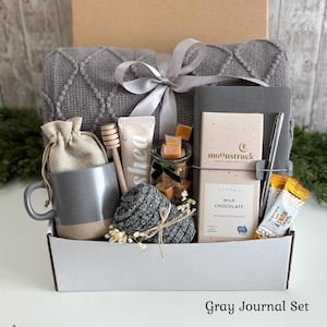 May include: A gift box containing a gray textured throw blanket with a silver ribbon, a gray and beige mug, a small burlap pouch, a wooden honey dipper, a tube of shea cream, a jar of caramels, a gray journal, a silver pen, and milk chocolate.