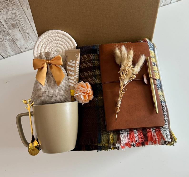 K&ouml;nnte beinhalten: Ge&ouml;ffnete Geschenkbox mit beigem Becher, goldenem L&ouml;ffel, Jutebeutel mit Schleife, Untersetzer, Honigtube, Kerze, Lederjournal, Stift und kariertem Schal. Die Box zeigt die enthaltenen Artikel.