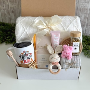 May include: A gift box containing a white textured blanket with a cream ribbon, a white mug with floral Mama text, a honey dipper, a glass straw, a Moonstruck milk chocolate bar, a rose hand cream, a crochet bunny rattle, a pink bear candle, and herbal tea.