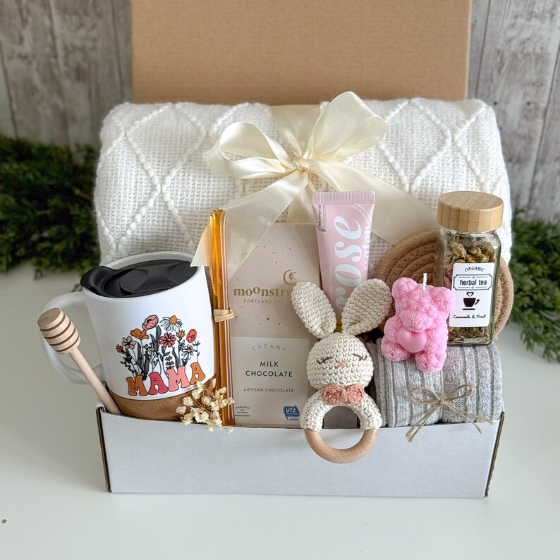 May include: A gift box containing a white textured blanket with a cream ribbon, a white mug with floral Mama text, a honey dipper, a glass straw, a Moonstruck milk chocolate bar, a rose hand cream, a crochet bunny rattle, a pink bear candle, and herbal tea.