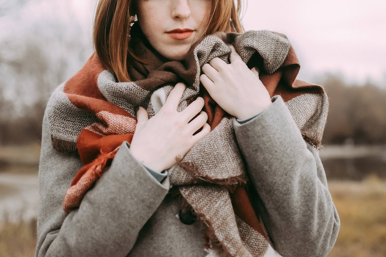 May include: A woman wearing a gray coat with a brown, red, and white patterned scarf wrapped around her neck.