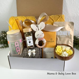 K&ouml;nnte beinhalten: Eine Geschenkbox mit einem Milchglasbecher mit der Aufschrift MAMA, einem Glas Kr&auml;utertee, einer geh&auml;kelten Hasenrassel, einer Tafel Milchschokolade, einem Honigbeutel und einer Holzschale mit sukkulentenf&ouml;rmigen Kerzen, gebunden mit einem beigen Band.