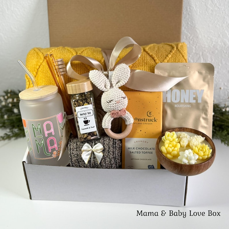 K&ouml;nnte beinhalten: Eine Geschenkbox mit einem Milchglasbecher mit der Aufschrift MAMA, einem Glas Kr&auml;utertee, einer geh&auml;kelten Hasenrassel, einer Tafel Milchschokolade, einem Honigbeutel und einer Holzschale mit sukkulentenf&ouml;rmigen Kerzen, gebunden mit einem beigen Band.