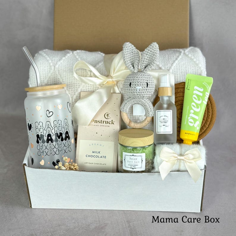 May include: A gift box containing a frosted glass tumbler with a bamboo lid and straw, a cream-colored knit blanket, a crochet bunny rattle, a Moonstruck milk chocolate bar, a jar of green bath salts, a spray bottle, and a green tea hand cream.